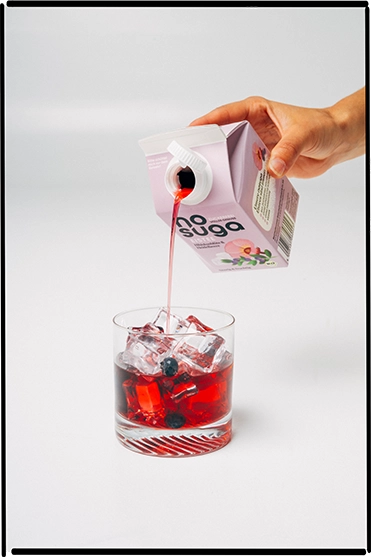Ein Getränkeglas gefüllt mit Eistee von nosuga vor weißem Hintergrund. Darüber ist eine Hand mit Getränkekarton und schenkt ein.