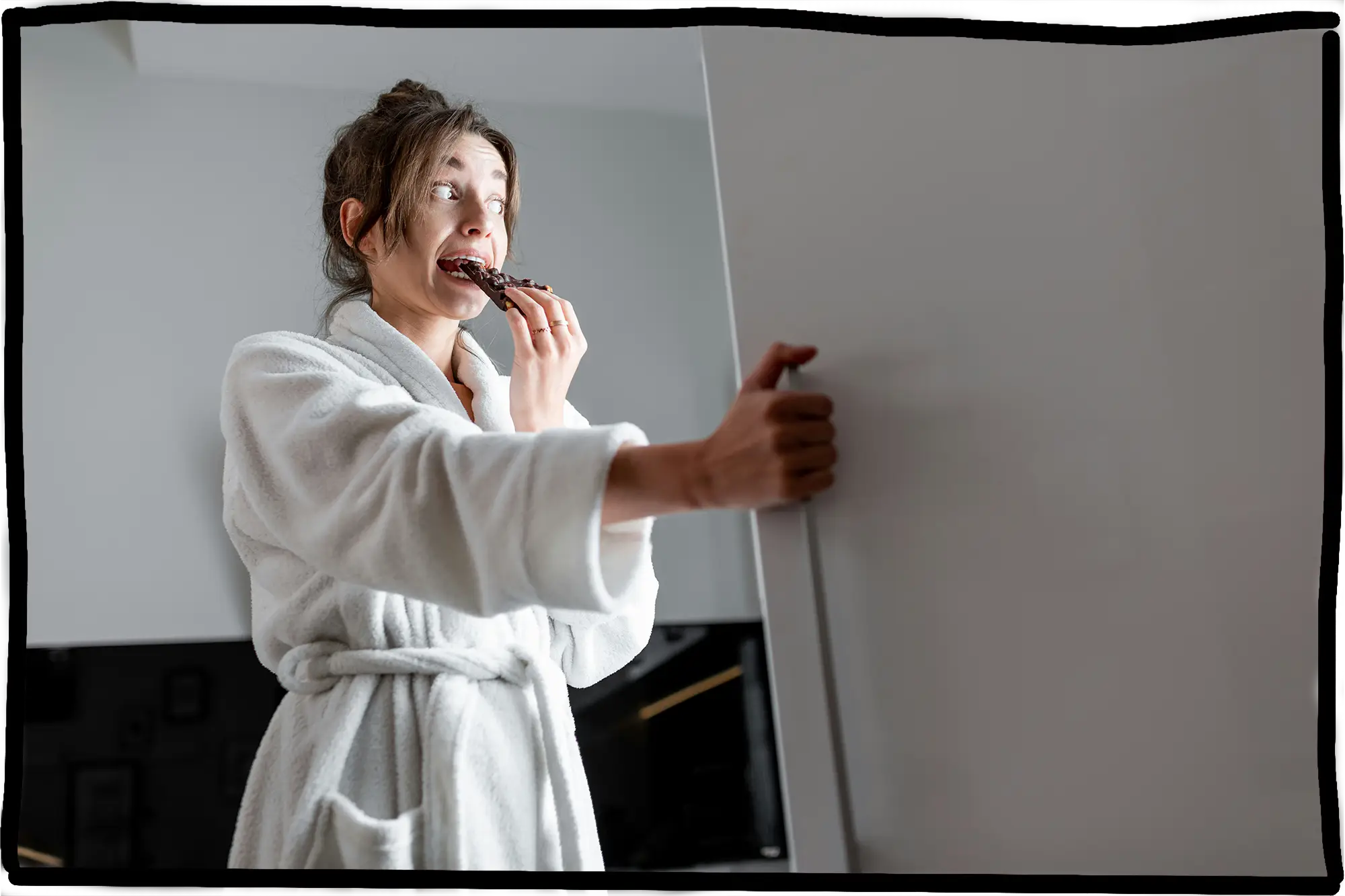 Eine Frau steht vor einem offenen Kühlschrank und beist ganz beschämt in eine Tafel Schokolade