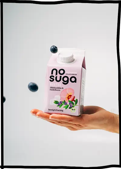 Eine Hand hält einen Getränkekarton von nosuga. Drum herum sind Blaubeeren.