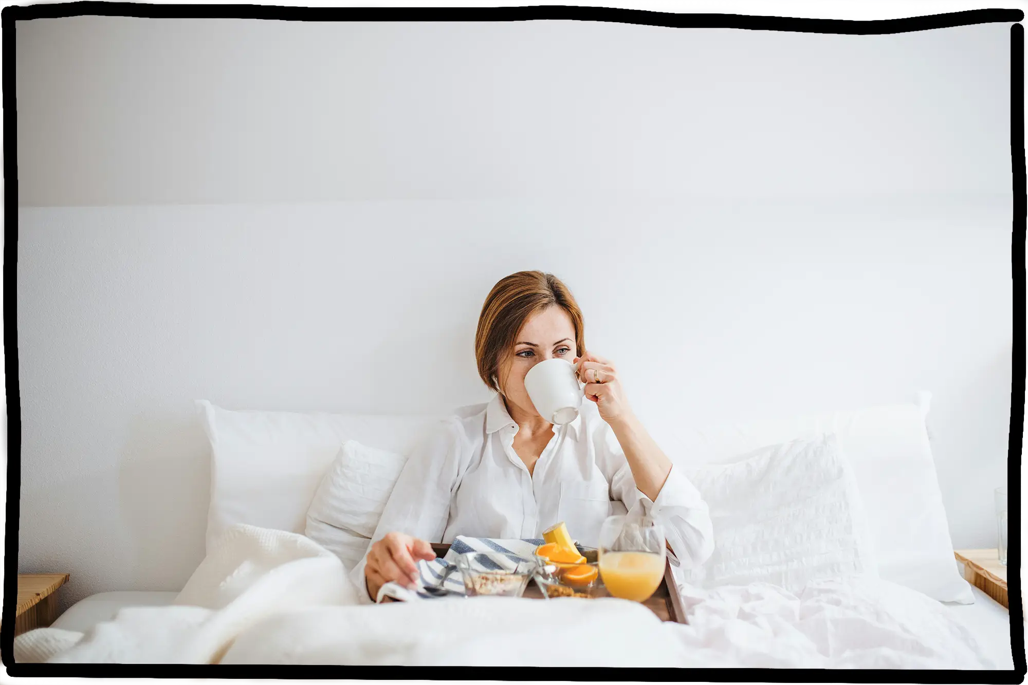 Eine Frau sitzt in einem weißen, gemütlichen Bett und trinkt aus einer weißen Tasse. Vor ihr steht ein Frühstückstablett mit Orangensaft, Obst und Müsli. Die Stimmung wirkt ruhig und entspannt.