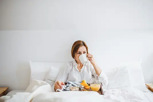 Eine Frau sitzt in einem weißen, gemütlichen Bett und trinkt aus einer weißen Tasse. Vor ihr steht ein Frühstückstablett mit Orangensaft, Obst und Müsli. Die Stimmung wirkt ruhig und entspannt.