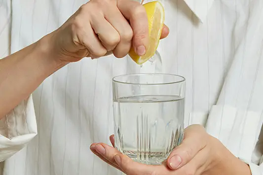 Eine Person presst eine Zitronenhälfte über ein Glas Wasser aus, das sie in der anderen Hand hält.