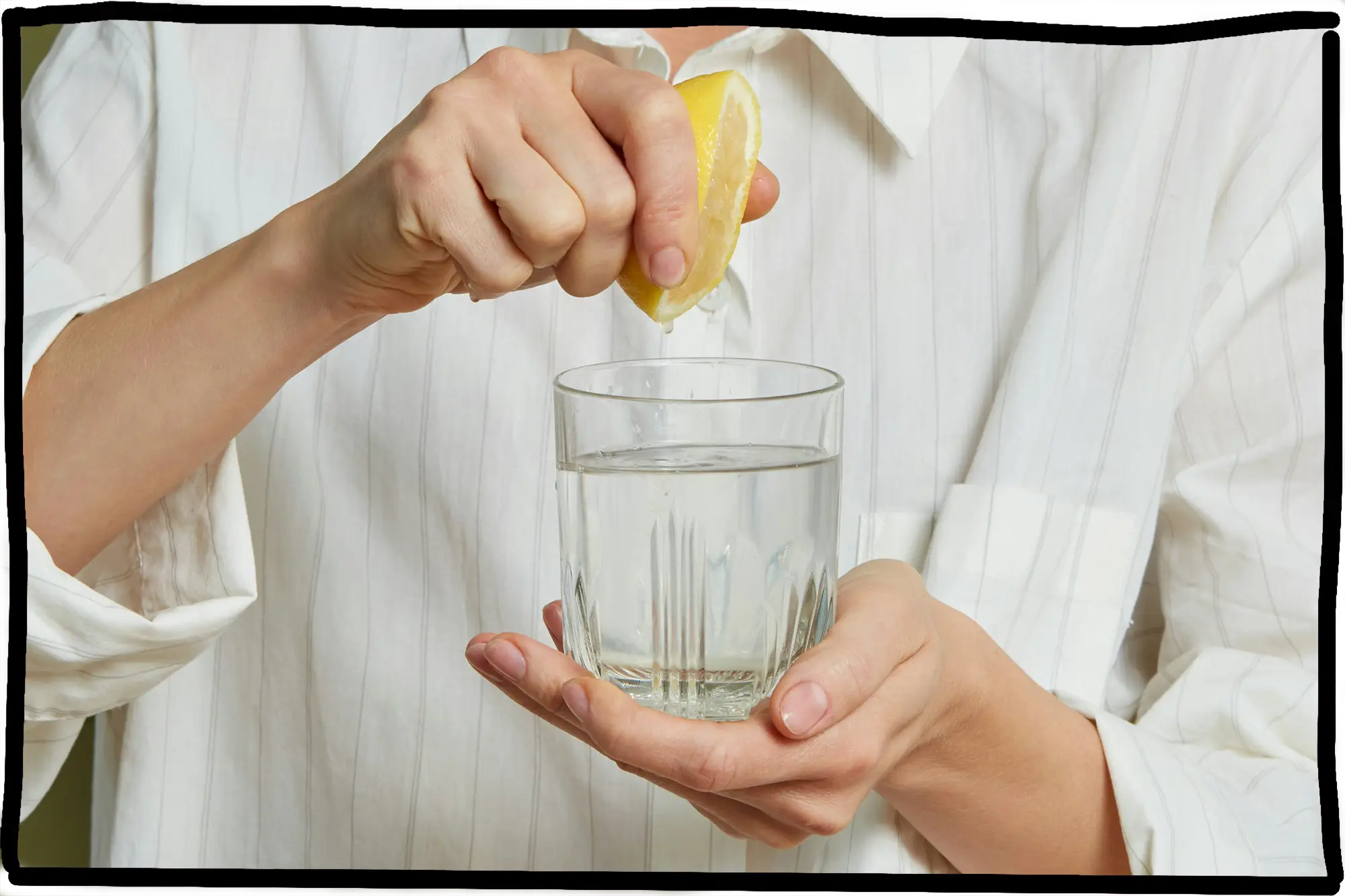 Eine Person presst eine Zitronenhälfte über ein Glas Wasser aus, das sie in der anderen Hand hält.