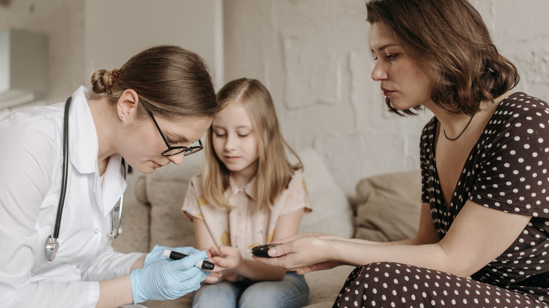 Blutzuckermessung bei einem Kind in einer häuslichen Umgebung. Eine Person mit Stethoskop und Schutzhandschuhen führt die Messung durch, während eine weitere Person daneben sitzt.